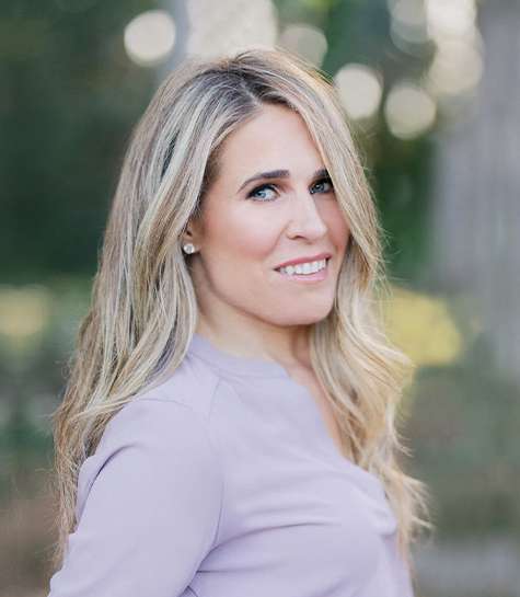 A woman with long, wavy blonde hair, wearing a light purple top, stands outdoors and smiles at the camera. The background is blurred with greenery and sunlight.