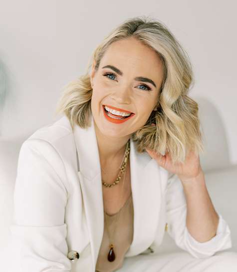 A smiling woman with blonde, wavy hair sits on a white couch. She wears a white blazer over a beige top, a gold necklace, and bright red lipstick, looking confidently at the camera.
