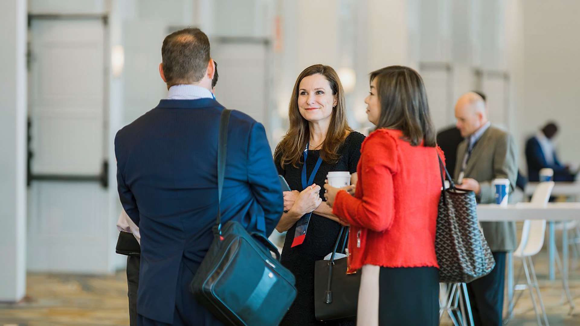 A group of professionals stand in conversation at a modern conference venue. One woman, holding a coffee cup and wearing a blue dress, smiles at a man in a suit. Others engage nearby, with tables and chairs in the background.