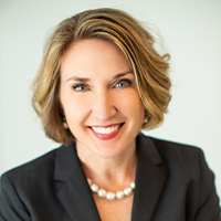 Smiling woman with short, light brown hair wearing a dark blazer, white top, and pearl necklace, pictured against a light, plain background.