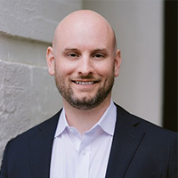 A bald man with a beard, wearing a dark suit jacket and white shirt, stands smiling in front of a white brick wall and doorway.