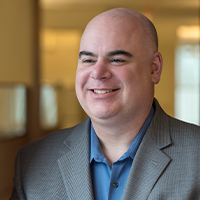 A smiling bald man wearing a gray suit jacket and blue collared shirt stands indoors with a softly blurred background.