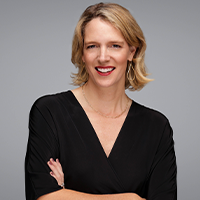 A woman with blonde hair, wearing a black V-neck top and gold hoop earrings, smiles confidently with her arms crossed against a plain gray background.