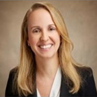 A woman with long blonde hair, wearing a black blazer over a white top, smiles at the camera against a neutral, light brown background.