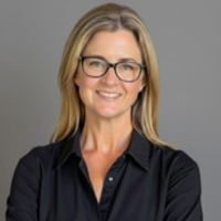 A woman with long blonde hair and glasses smiles at the camera. She is wearing a black button-up shirt and stands in front of a plain gray background.