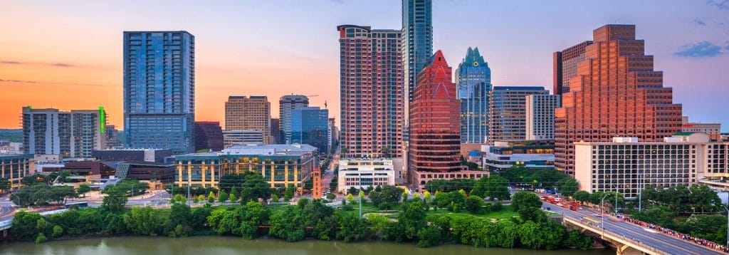 Downtown Austin skyline at sunset, featuring tall modern buildings with glass facades, colorful lights, and a river with green trees in the foreground.