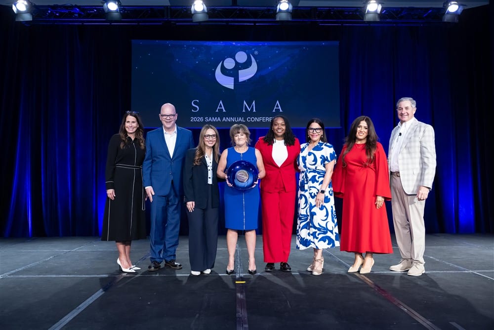 Eight people stand on stage at the SAMA 2024 Annual Conference, with a woman in the center holding an award. They are smiling and dressed in business or formal attire, with a dark blue curtain and SAMA logo in the background.
