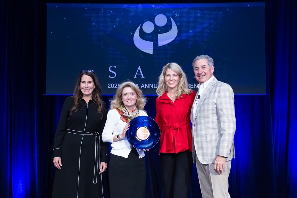 Four people stand on stage at an awards event; one woman holds a blue glass award plate. A large event sign and blue curtains are in the background. All are smiling and dressed in business or semi-formal attire.