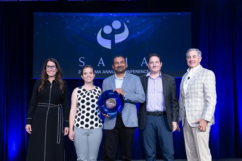 Five people stand on stage at a conference, one holding a blue glass award. They are smiling in front of a large screen displaying the SAIA logo and conference information, with blue and purple lighting in the background.