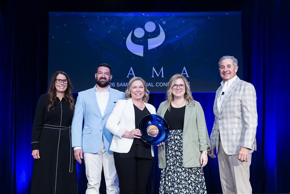 Five people stand on stage during an award ceremony. The woman in the center holds a blue glass award. They are dressed in business attire, with a blue and black conference backdrop behind them displaying the AAMA logo.