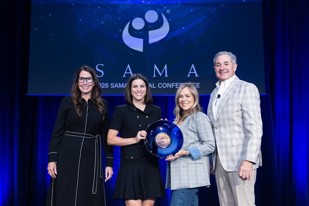 Four people stand on a stage at the SAMA 2026 Annual Conference. Two women in the center hold a blue award plate, with a large SAMA logo and conference banner in the background.