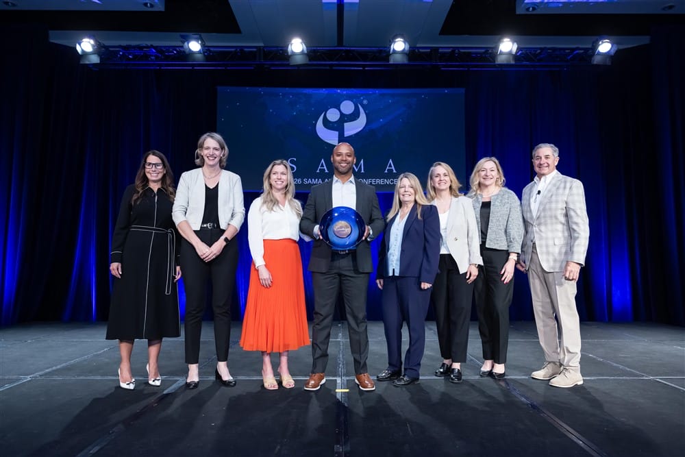 A group of eight people, dressed in business attire, stand on stage smiling. The person in the center holds a blue glass award. A conference logo is projected on a screen behind them.