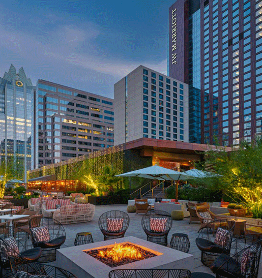 A stylish rooftop patio at dusk features a central firepit, modern lounge seating, string lights, lush greenery, and tall city buildings, including the JW Marriott, in the background.