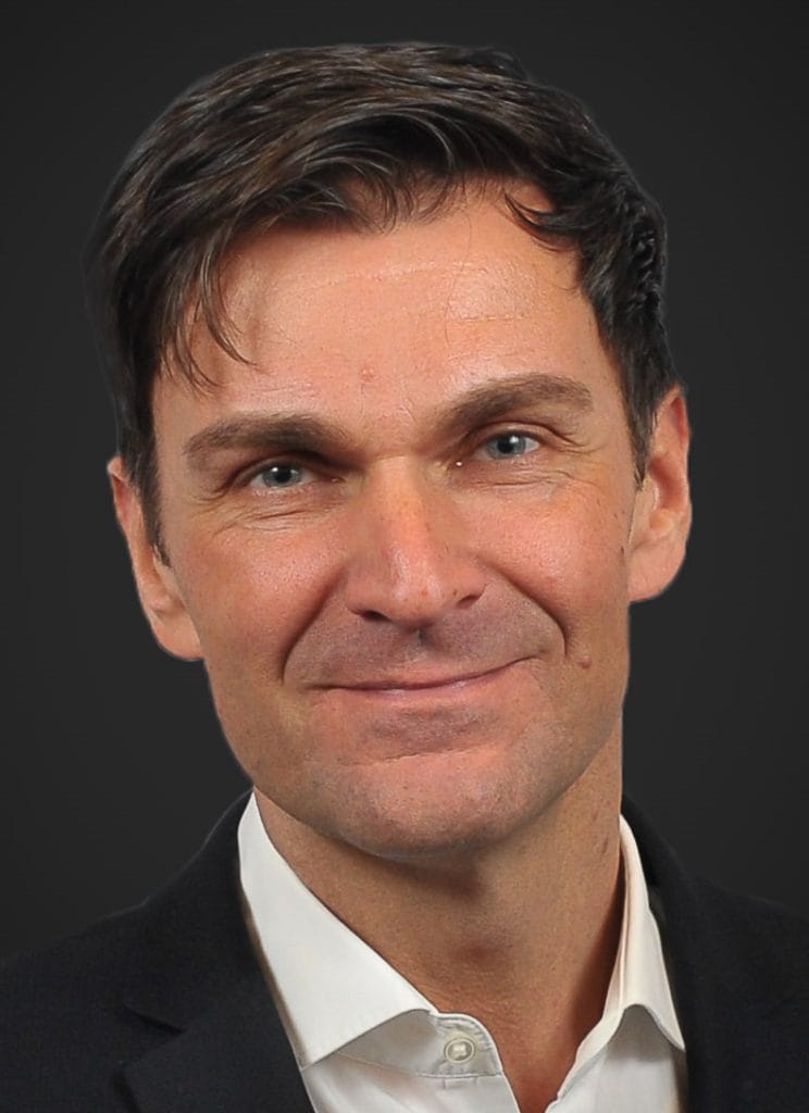 A man with short brown hair, blue eyes, and light skin is wearing a dark suit jacket and a white shirt, smiling slightly in front of a plain dark background.
