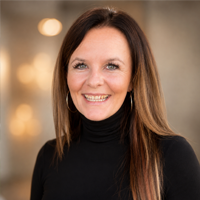 Woman with long brown hair wearing a black turtleneck smiles at the camera. The background is softly blurred with warm lighting.