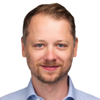 Smiling man with short light brown hair and light facial hair, wearing a light blue collared shirt, facing forward against a plain white background.