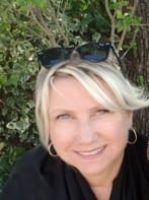 A smiling woman with short blonde hair and sunglasses on her head is outdoors in front of green foliage.