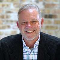 A middle-aged man with short gray hair and a beard is smiling, wearing a dark jacket over a plaid shirt. The background features a blurred brick wall.
