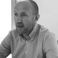 Black and white photo of a man with a beard, wearing a checkered shirt, sitting at a table and looking slightly to the side with a neutral expression.