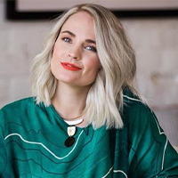 A woman with shoulder-length blonde hair and bright red lipstick is wearing a green patterned top and a necklace with circular pendants, smiling slightly while sitting indoors.