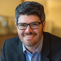 A smiling man with short dark hair and a beard, wearing glasses, a dark blazer, and a light blue shirt, sits indoors with a blurred warm-toned background.