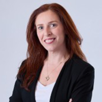 A woman with long red hair, wearing a black blazer over a white top, stands against a light background, smiling with her arms crossed.