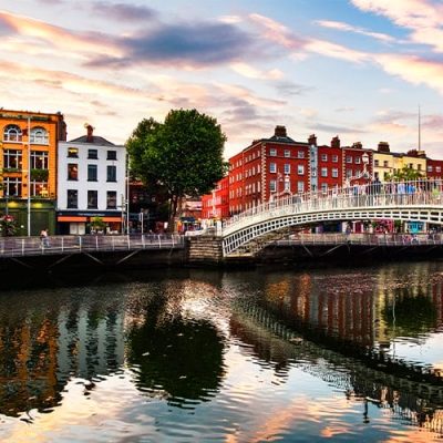 A white pedestrian bridge crosses a river lined with colorful buildings, trees, and people, reflecting in the calm water under a partly cloudy sky at sunset.