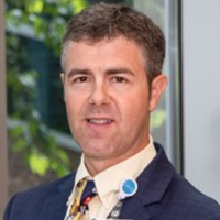 A man with short brown hair wearing a dark suit, light shirt, patterned tie, and a lanyard stands indoors near a window with greenery visible outside.
