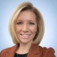 A woman with straight blonde hair, wearing a brown blazer over a black top, smiles at the camera against a light blue background.