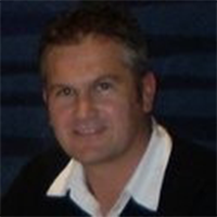 A man with short, dark hair is smiling at the camera. He is wearing a white collared shirt under a dark sweater, with a dark blue background behind him.