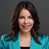 A woman with long dark hair, wearing a teal blazer over a black top and layered gold necklaces, smiles at the camera against a plain dark gray background.