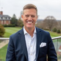 Smiling man in a navy suit and white shirt stands outside, with a green lawn, trees, and a brick building in the background on a cloudy day.