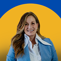 A woman with long, wavy hair wearing a blue blazer and white collared shirt smiles in front of a blue and yellow circular background.