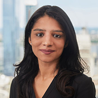 A woman with long dark hair, wearing a black top, stands in front of a window with a blurred cityscape in the background. She is smiling slightly and looking at the camera.