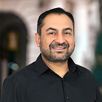 Smiling man with short black hair and a beard, wearing a black collared shirt, standing in front of a blurred outdoor background.