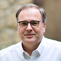 A middle-aged man with short brown hair, glasses, and a white button-down shirt is smiling softly. The background appears to be outdoors with a blurred stone wall.